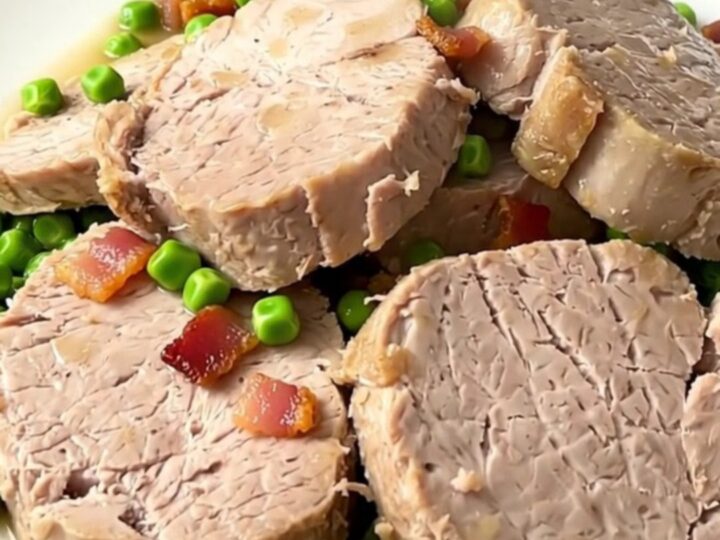A close-up of sliced pork tenderloin medallions topped with green peas and bacon lardons on a white plate.