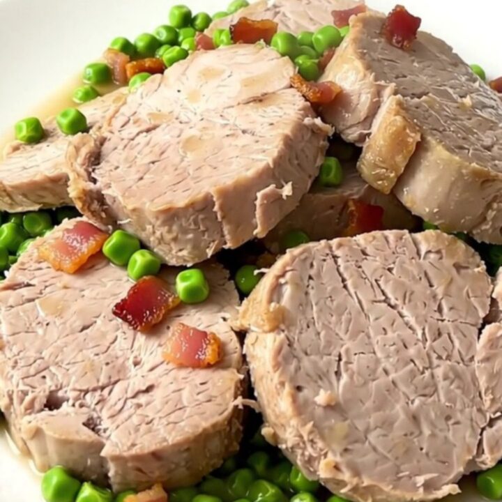 A close-up of sliced pork tenderloin medallions topped with green peas and bacon lardons on a white plate.