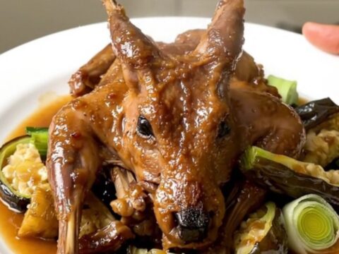A white plate featuring tender braised rabbit pieces surrounded by glazed carrots, leeks, and onions in a light cider sauce.