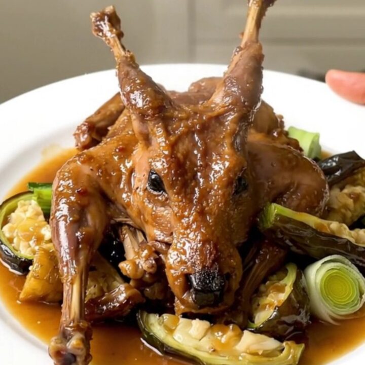 A white plate featuring tender braised rabbit pieces surrounded by glazed carrots, leeks, and onions in a light cider sauce.