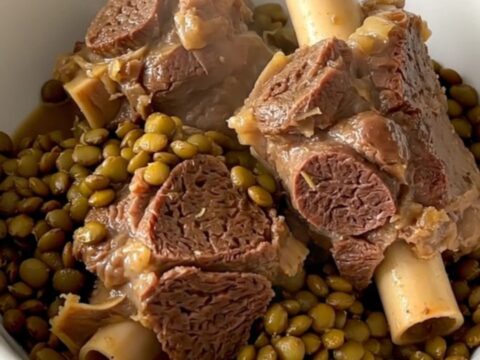 A steaming bowl of tender braised lamb neck slices served over a bed of glossy, cooked green lentils in a bright kitchen.