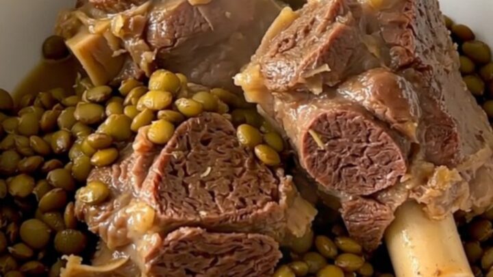 A steaming bowl of tender braised lamb neck slices served over a bed of glossy, cooked green lentils in a bright kitchen.
