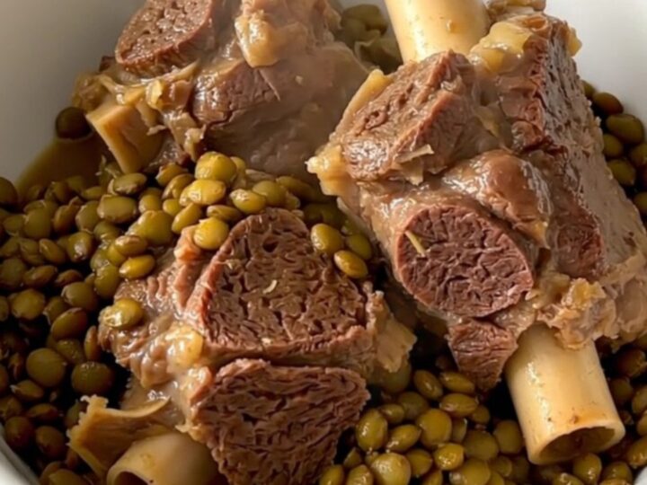 A steaming bowl of tender braised lamb neck slices served over a bed of glossy, cooked green lentils in a bright kitchen.