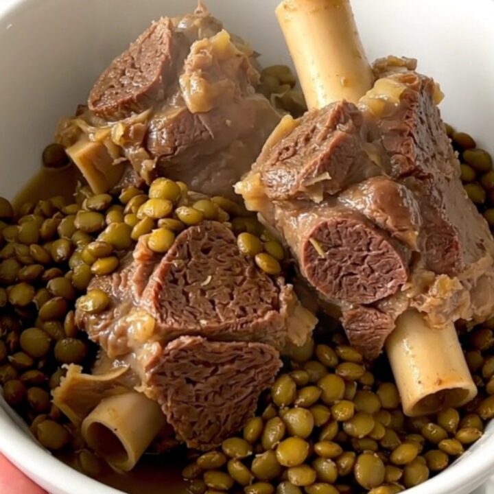 A steaming bowl of tender braised lamb neck slices served over a bed of glossy, cooked green lentils in a bright kitchen.