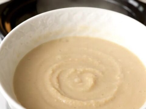 A white bowl filled with smooth, creamy, pale-colored celeriac and apple soup, steaming slightly.