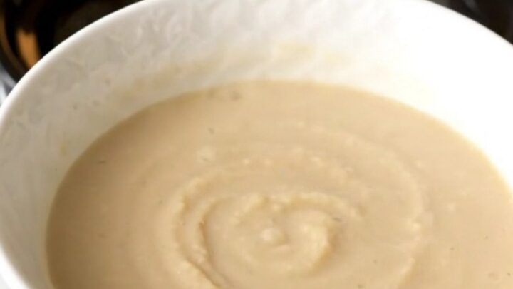 A white bowl filled with smooth, creamy, pale-colored celeriac and apple soup, steaming slightly.
