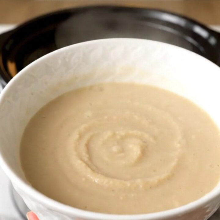 A white bowl filled with smooth, creamy, pale-colored celeriac and apple soup, steaming slightly.