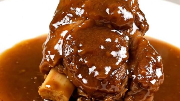 A close-up of a tender, slow-cooked lamb shank resting on a white plate with a glossy, dark amber sauce.