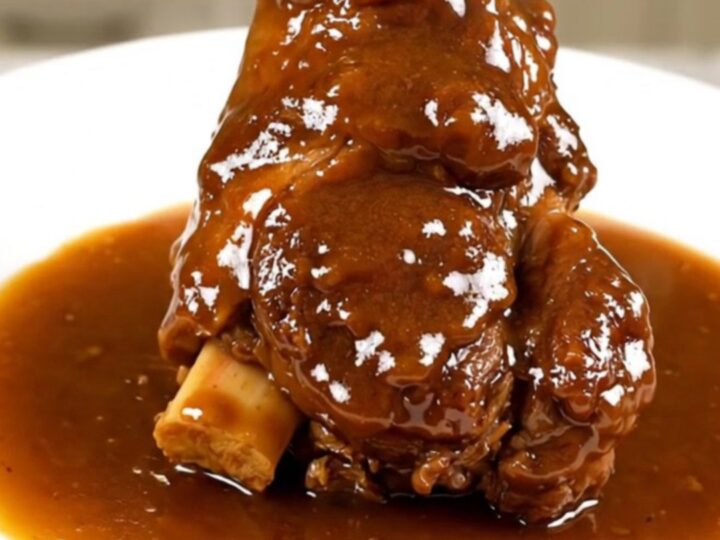 A close-up of a tender, slow-cooked lamb shank resting on a white plate with a glossy, dark amber sauce.
