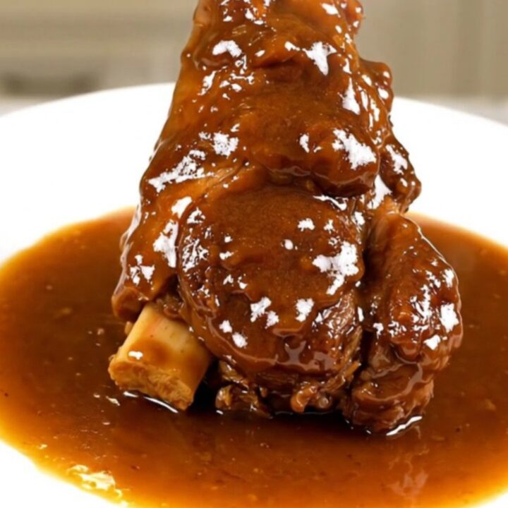 A close-up of a tender, slow-cooked lamb shank resting on a white plate with a glossy, dark amber sauce.