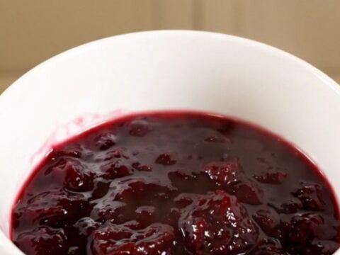 A white bowl filled with glossy, deep purple plum jam, held in a hand with blurred kitchen cabinets in the background.