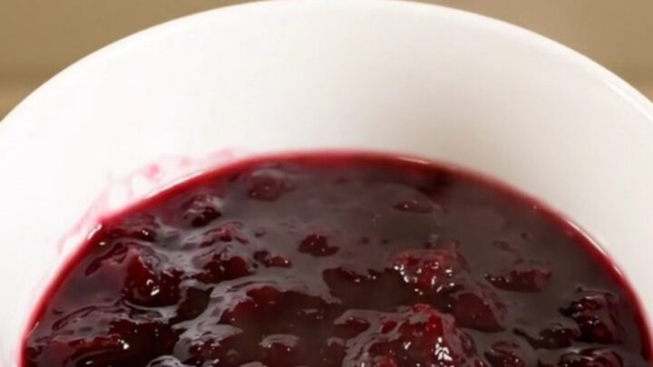 A white bowl filled with glossy, deep purple plum jam, held in a hand with blurred kitchen cabinets in the background.