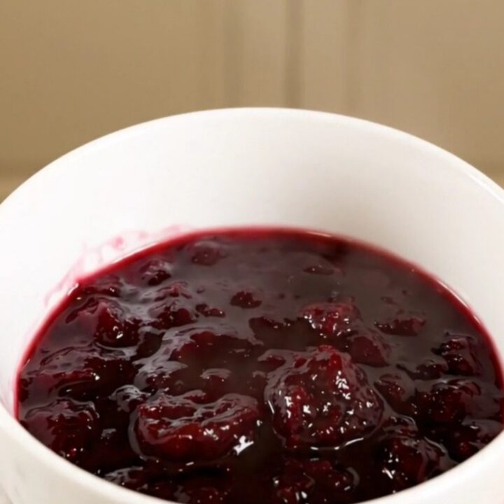 A white bowl filled with glossy, deep purple plum jam, held in a hand with blurred kitchen cabinets in the background.