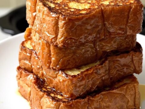 A close-up of golden, glossy brioche French toast served on a white plate in a bright kitchen.
