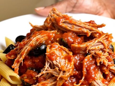 A white plate topped with shredded pork in a rich tomato and olive sauce over penne pasta.