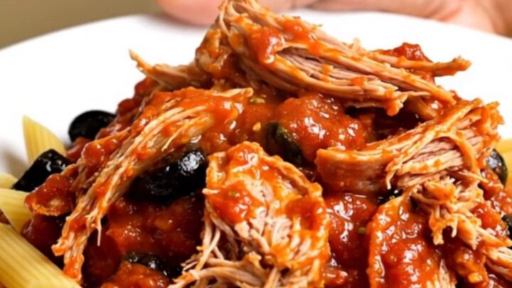 A white plate topped with shredded pork in a rich tomato and olive sauce over penne pasta.