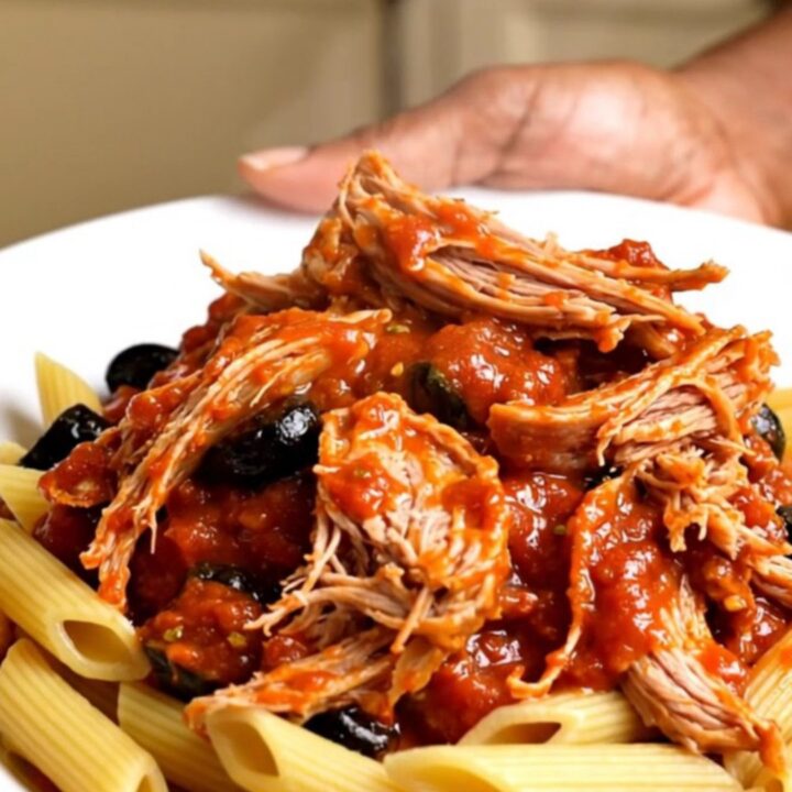 A white plate topped with shredded pork in a rich tomato and olive sauce over penne pasta.