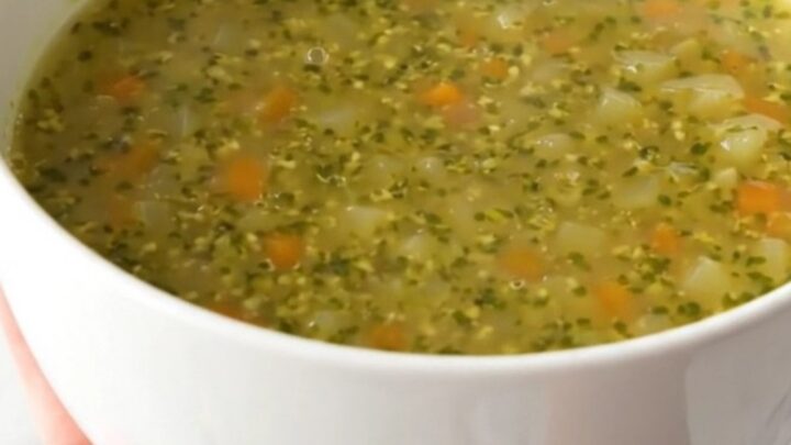 A white ceramic bowl filled with steaming, glossy autumn pistou soup, held by a hand against a blurred kitchen background.