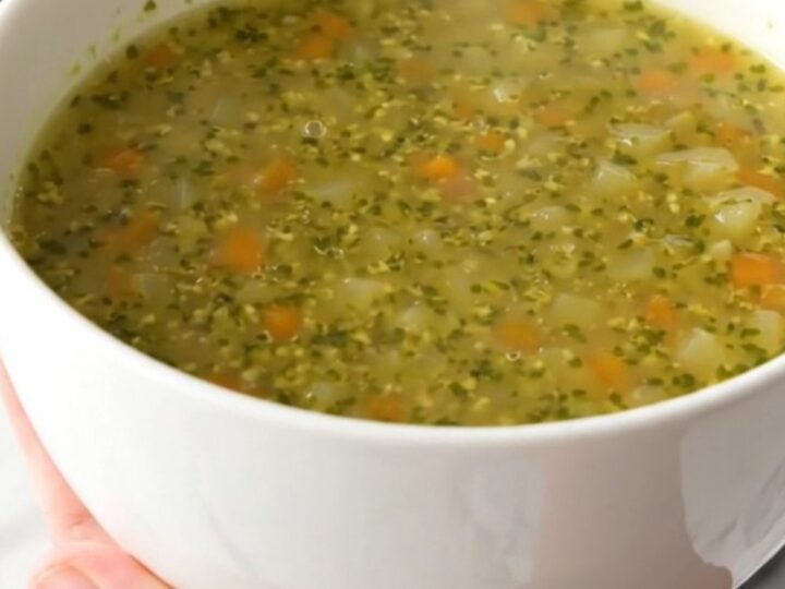 A white ceramic bowl filled with steaming, glossy autumn pistou soup, held by a hand against a blurred kitchen background.