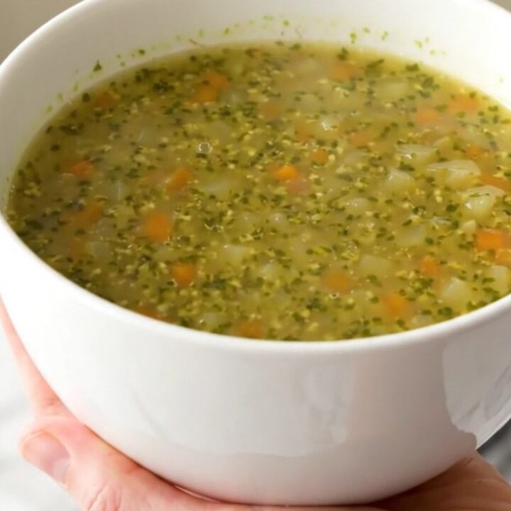 A white ceramic bowl filled with steaming, glossy autumn pistou soup, held by a hand against a blurred kitchen background.