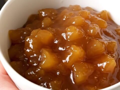 A white bowl filled with glossy, spiced applesauce, garnished with a cinnamon stick, against a blurred kitchen background.