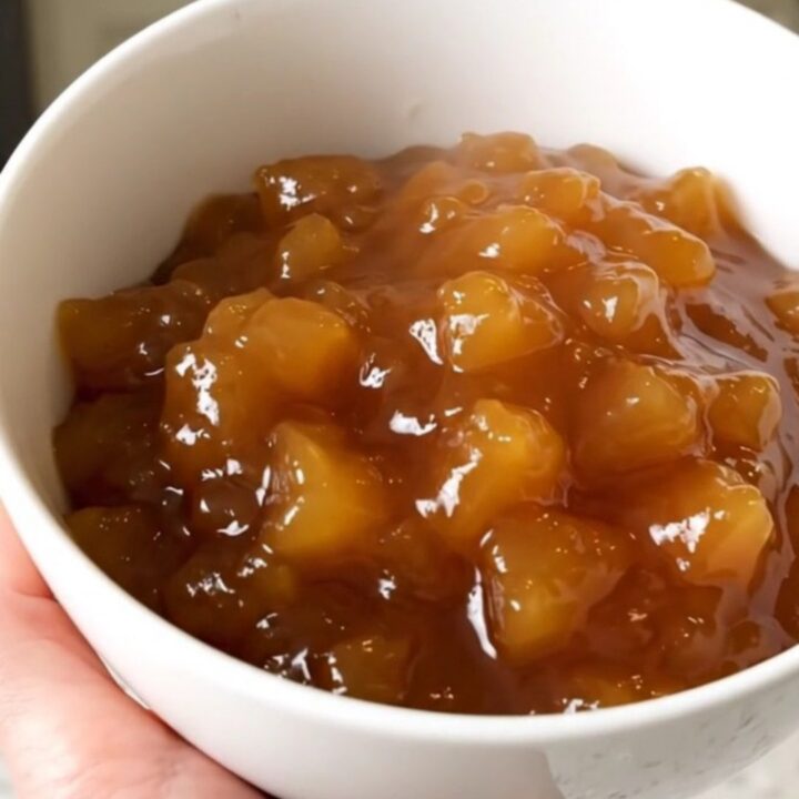 A white bowl filled with glossy, spiced applesauce, garnished with a cinnamon stick, against a blurred kitchen background.