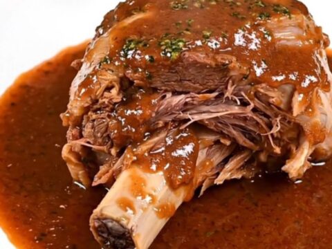 A close-up shot of a tender veal osso buco shank served on a white plate with a rich red wine sauce and vegetables.