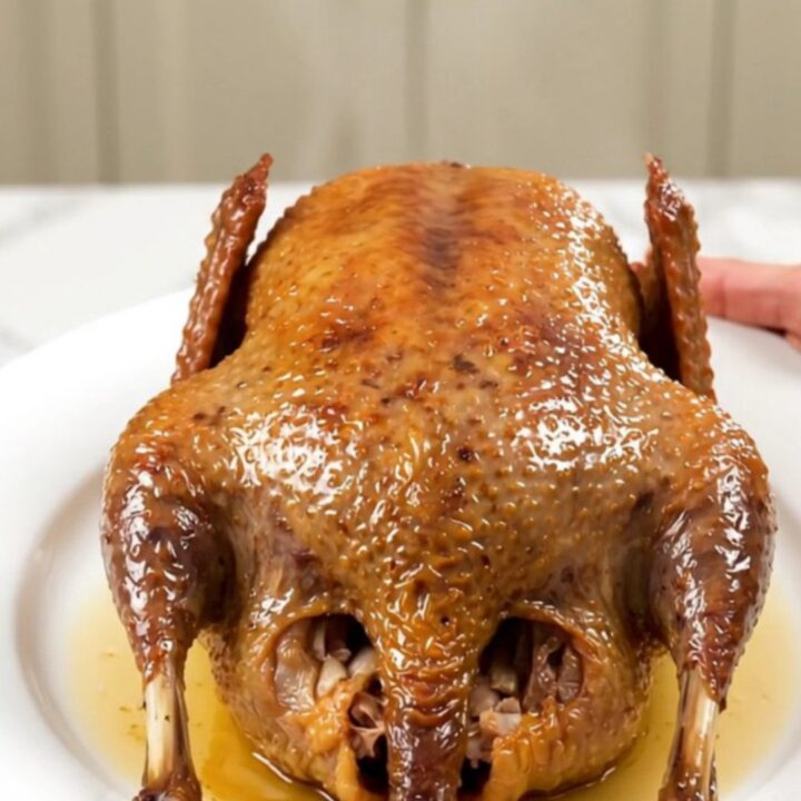 A whole, golden-roasted guinea fowl resting on a white plate with fresh thyme and caramelized garlic.