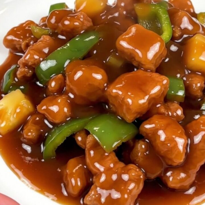 A close-up of glossy sweet and sour pork cubes with pineapple and green peppers in a slow cooker.