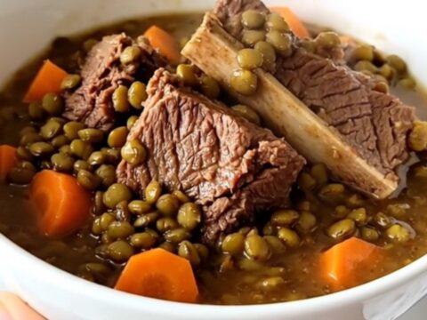 A steaming bowl of beef shank and lentil stew with visible carrot slices and herbs on a wooden table.