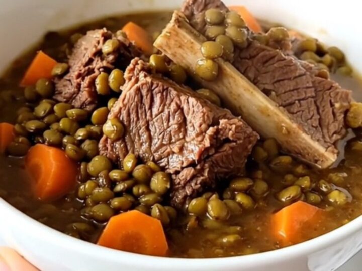 A steaming bowl of beef shank and lentil stew with visible carrot slices and herbs on a wooden table.
