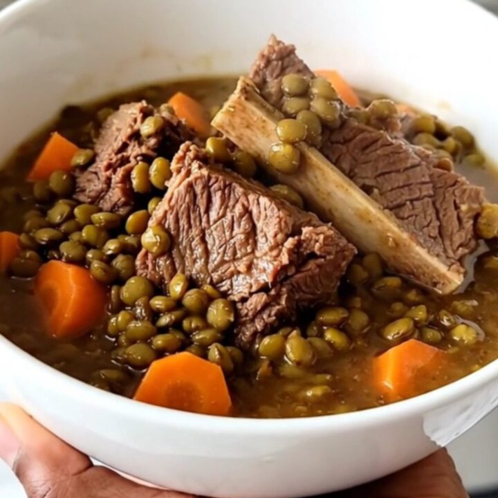 A steaming bowl of beef shank and lentil stew with visible carrot slices and herbs on a wooden table.
