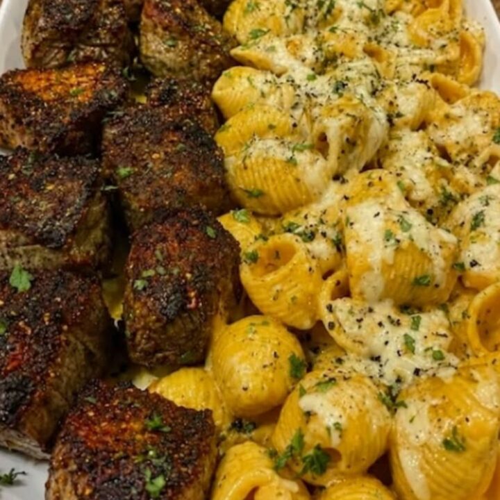 A large serving platter featuring golden cheesy conchiglie pasta shells and seared steak bites under warm lighting.