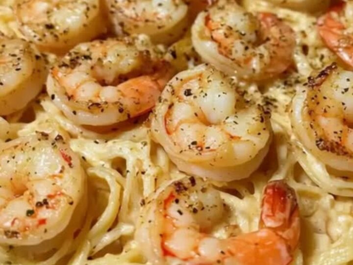 A close-up of creamy linguine topped with golden jumbo shrimp in a foil pan.