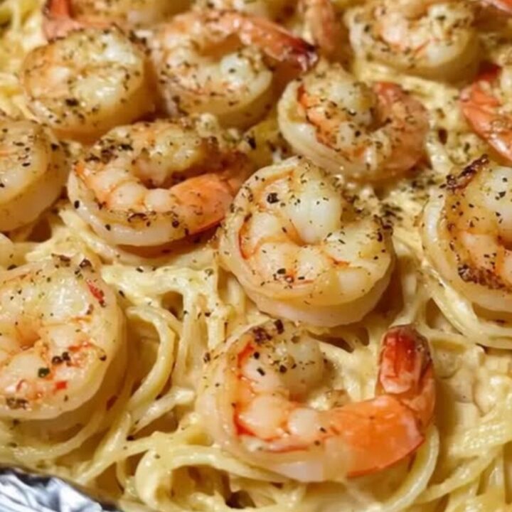 A close-up of creamy linguine topped with golden jumbo shrimp in a foil pan.