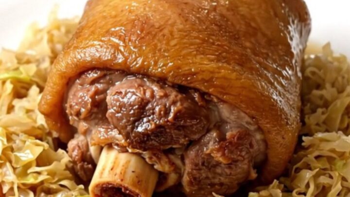 A glossy, tender pork shank nestled in a bed of warm sauerkraut on a white plate.