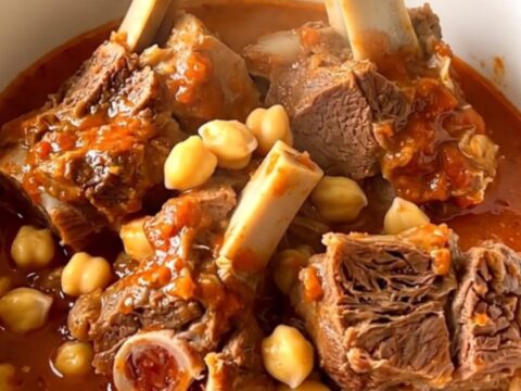 A steaming bowl of lamb neck stew with chickpeas in a rich tomato sauce against a cream-colored kitchen background.