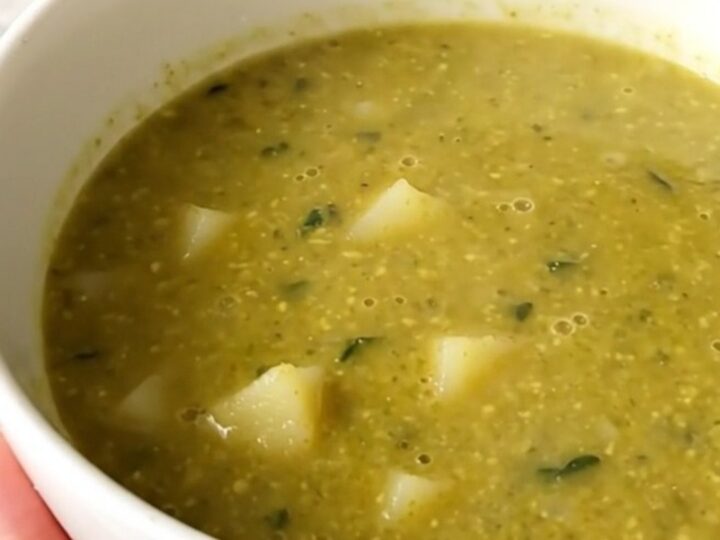 A steaming, glossy bowl of vibrant green spinach and potato soup held by a hand in a bright kitchen.