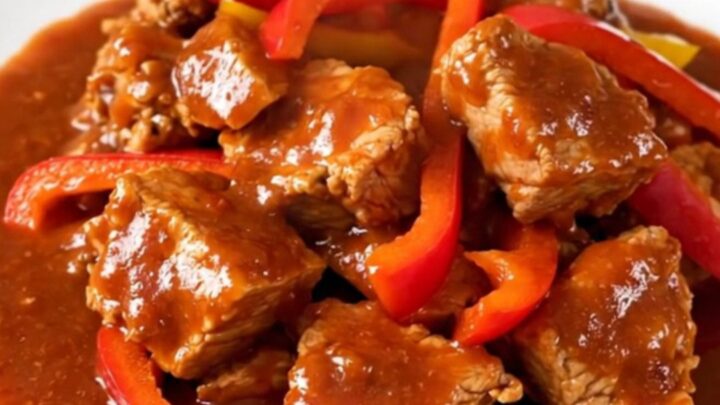 A white round plate filled with tender slow-cooked pork cubes and colorful bell peppers in a rich red sauce.
