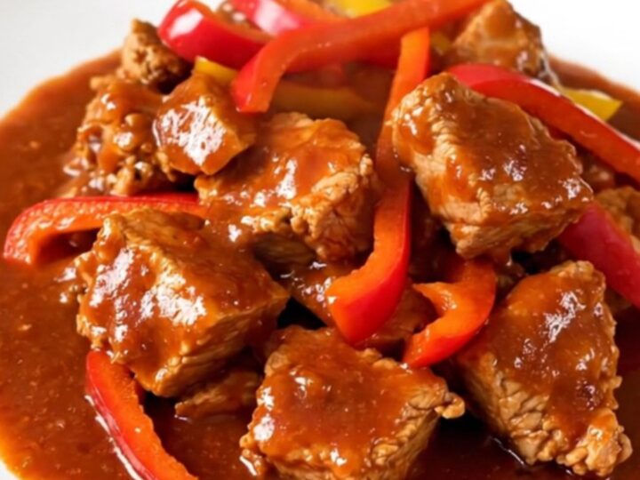 A white round plate filled with tender slow-cooked pork cubes and colorful bell peppers in a rich red sauce.