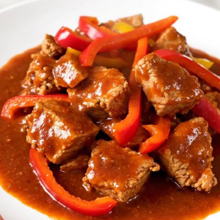 A white round plate filled with tender slow-cooked pork cubes and colorful bell peppers in a rich red sauce.