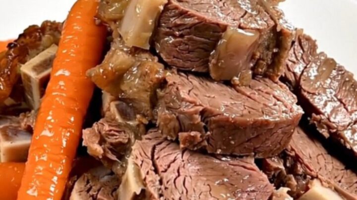 A slow-cooked boneless leg of lamb served on a white plate with tender carrots and shallots, glistening with pan juices.