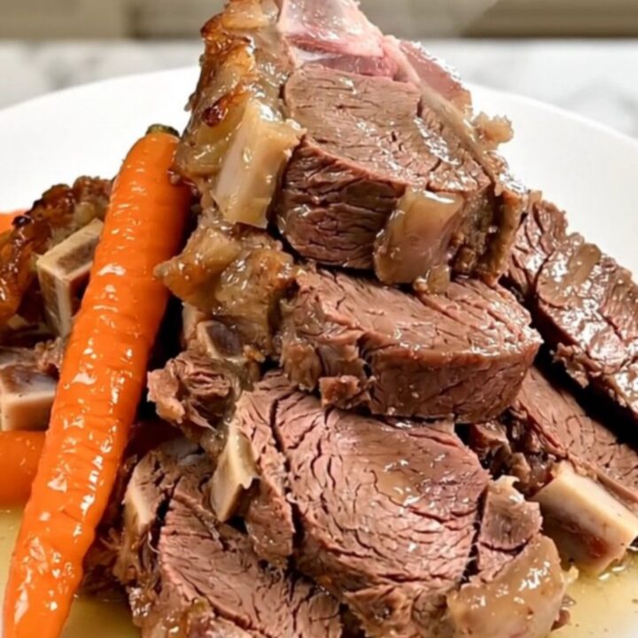 A slow-cooked boneless leg of lamb served on a white plate with tender carrots and shallots, glistening with pan juices.
