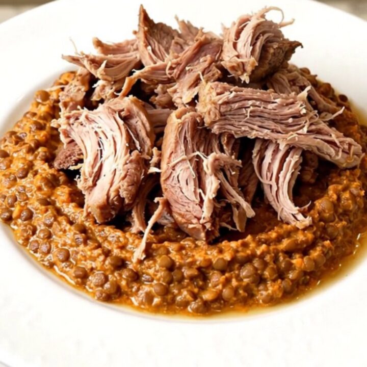 A close-up of tender, shredded pork shoulder served with a creamy red lentil and carrot puree on a white plate.