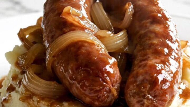 A black slow cooker filled with raw Toulouse sausages and sliced sweet onions on a white speckled quartz countertop.