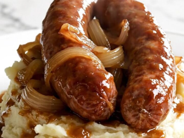 A black slow cooker filled with raw Toulouse sausages and sliced sweet onions on a white speckled quartz countertop.