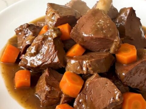 A close-up of a bowl of slow-cooked, tender beer-braised veal stew with carrots and onions in a rich, glossy brown sauce.