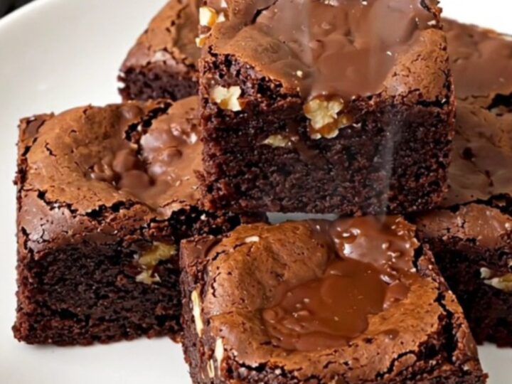 A close-up of a warm, fudgy walnut brownie square on a white plate, showing a dense and glossy texture.