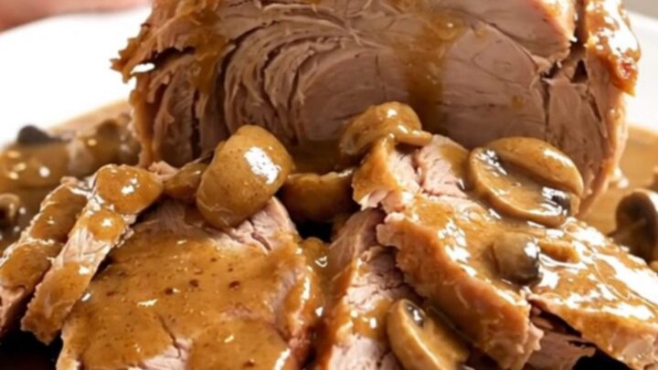 A slow-cooked pork roast with creamy mushroom sauce in a slow cooker on a white speckled countertop.