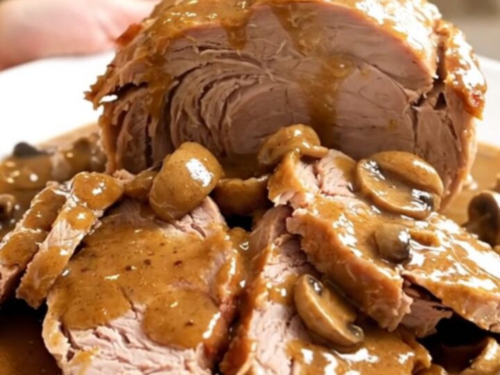 A slow-cooked pork roast with creamy mushroom sauce in a slow cooker on a white speckled countertop.